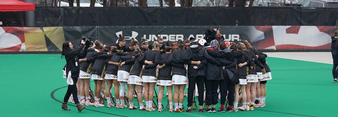 A group of athletes is gathered in a huddle on a sports field, wearing matching uniforms and gear. The ground is green, and there is signage with a brand logo in the background. Trees and parked cars can be seen behind a fence surrounding the field. A person on the left side is capturing the moment with a camera.