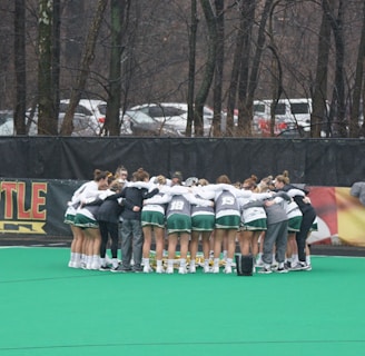 A group of athletes huddled together in a circle on a green sports field. They are wearing uniforms with green shorts and white tops, indicating a team sport environment. Lacrosse sticks are lined up against a fence in the background. Trees and parked cars are visible beyond the fence, suggesting an outdoor location.