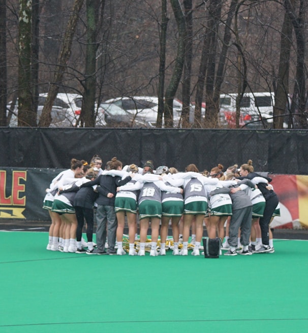 A group of athletes huddled together in a circle on a green sports field. They are wearing uniforms with green shorts and white tops, indicating a team sport environment. Lacrosse sticks are lined up against a fence in the background. Trees and parked cars are visible beyond the fence, suggesting an outdoor location.