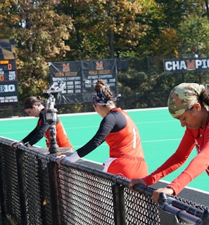 A group of athletes wearing orange uniforms are on the sideline of a sports field, resting against a fence. Behind them, a large digital scoreboard displays team names and scores. The field is bright green, and several banners with sponsor logos and championship wins are visible. The athletes appear to be taking a break, possibly before or during a field hockey game.