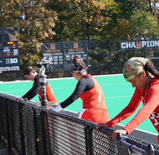 A group of athletes wearing orange uniforms are on the sideline of a sports field, resting against a fence. Behind them, a large digital scoreboard displays team names and scores. The field is bright green, and several banners with sponsor logos and championship wins are visible. The athletes appear to be taking a break, possibly before or during a field hockey game.