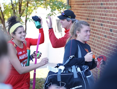A group of athletes in sports attire appears to be in a joyful mood. One person is holding a field hockey stick and wearing a headband, while another carries a sports bag. They are gathered near a brick wall with trees in the background, sharing a light moment, likely after a sports event.