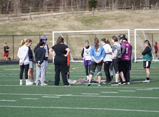A group of people gathers on a sports field, likely for a team discussion or practice session. They are dressed in athletic clothing, and some are holding or standing near lacrosse sticks. The setting appears to be an outdoor sports complex with a netted goal visible in the background. The field markings indicate it is a standard sports field, and the weather looks cool as some individuals are wearing long sleeves.