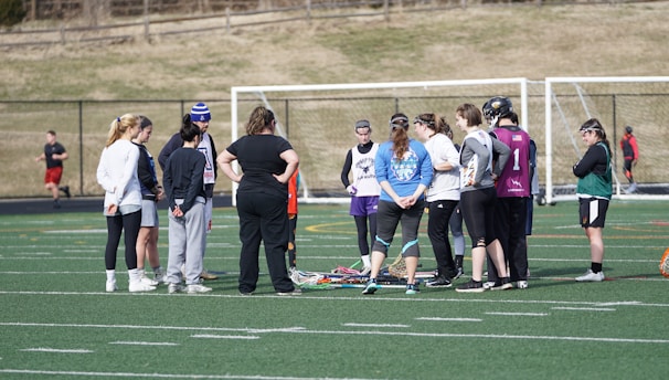 A group of people gathers on a sports field, likely for a team discussion or practice session. They are dressed in athletic clothing, and some are holding or standing near lacrosse sticks. The setting appears to be an outdoor sports complex with a netted goal visible in the background. The field markings indicate it is a standard sports field, and the weather looks cool as some individuals are wearing long sleeves.