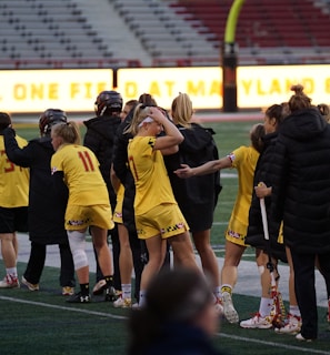 A group of athletes wearing yellow sports uniforms are standing on a sports field. Some are holding lacrosse sticks, and they appear to be interacting casually. Several people are dressed in black coats. In the background, there is a bright sign with text and parts of a stadium are visible.