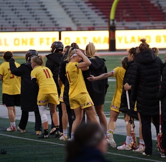 A group of athletes wearing yellow sports uniforms are standing on a sports field. Some are holding lacrosse sticks, and they appear to be interacting casually. Several people are dressed in black coats. In the background, there is a bright sign with text and parts of a stadium are visible.