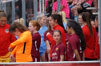 A group of young female athletes in sports attire are gathered together, some wearing red jackets or maroon jerseys with the logo of a soccer academy. They appear to be standing in front of a wire fence with spectators in the background, dressed in casual clothing.
