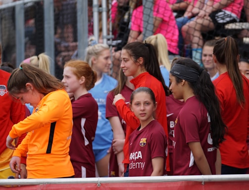 A group of young female athletes in sports attire are gathered together, some wearing red jackets or maroon jerseys with the logo of a soccer academy. They appear to be standing in front of a wire fence with spectators in the background, dressed in casual clothing.