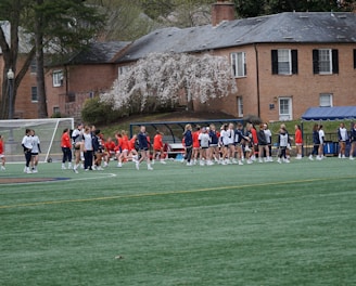 A group of people, primarily young athletes, are gathered on a sports field, some dressed in uniforms consisting of navy blue and red colors. In the background, there are several red-brick buildings and blossoming trees, suggesting a springtime setting. A goalpost can be seen, indicating that this is likely a game of soccer or lacrosse. The scene is lively and bustling with activity.