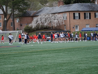 A group of people, primarily young athletes, are gathered on a sports field, some dressed in uniforms consisting of navy blue and red colors. In the background, there are several red-brick buildings and blossoming trees, suggesting a springtime setting. A goalpost can be seen, indicating that this is likely a game of soccer or lacrosse. The scene is lively and bustling with activity.