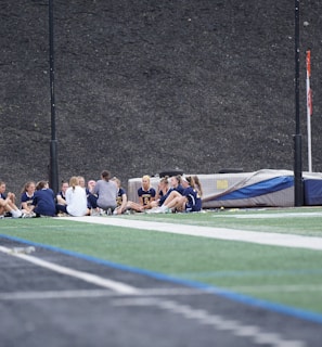 A group of people, likely a sports team, is sitting together on a field. They are wearing matching athletic uniforms, suggesting team unity, and seem to be engaged in a discussion or meeting. In the background, there is sports equipment and a high fence.