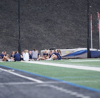 A group of people, likely a sports team, is sitting together on a field. They are wearing matching athletic uniforms, suggesting team unity, and seem to be engaged in a discussion or meeting. In the background, there is sports equipment and a high fence.