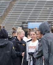 A group of people, likely athletes, are gathered together on a sports field. They are wearing black jackets with some individuals dressed in white sports uniforms underneath. A few people have their hair tied up in buns, and most are engaged in conversation. The background shows tiered stadium seats, suggesting the setting is an outdoor sports venue.