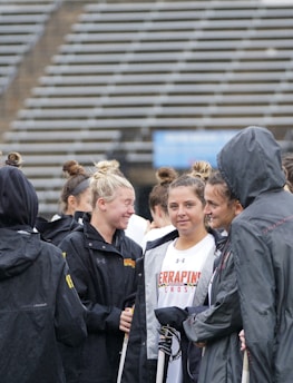 A group of people, likely athletes, are gathered together on a sports field. They are wearing black jackets with some individuals dressed in white sports uniforms underneath. A few people have their hair tied up in buns, and most are engaged in conversation. The background shows tiered stadium seats, suggesting the setting is an outdoor sports venue.
