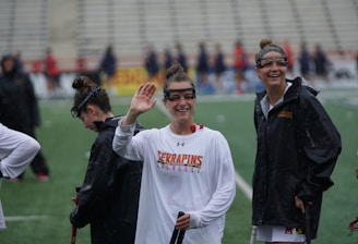 Several athletes in sports attire are on an outdoor sports field, interacting and smiling. One individual in the foreground waves with a friendly expression, while another stands beside her, also smiling. They wear protective goggles and gear associated with lacrosse. The background features indistinct figures in dark clothing against a backdrop of stadium seating.