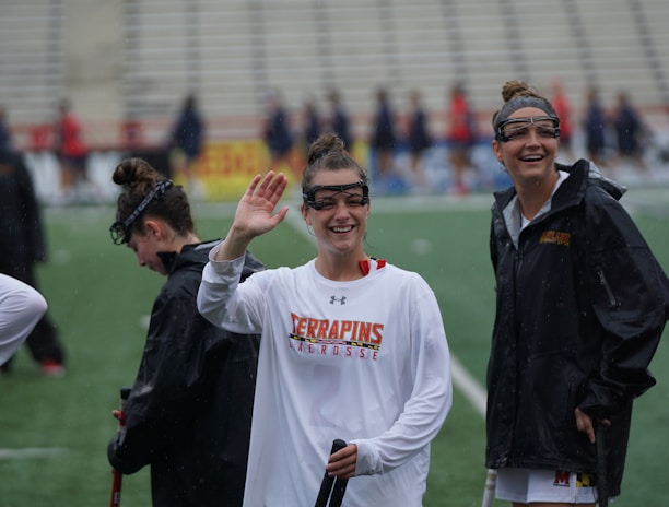 Several athletes in sports attire are on an outdoor sports field, interacting and smiling. One individual in the foreground waves with a friendly expression, while another stands beside her, also smiling. They wear protective goggles and gear associated with lacrosse. The background features indistinct figures in dark clothing against a backdrop of stadium seating.