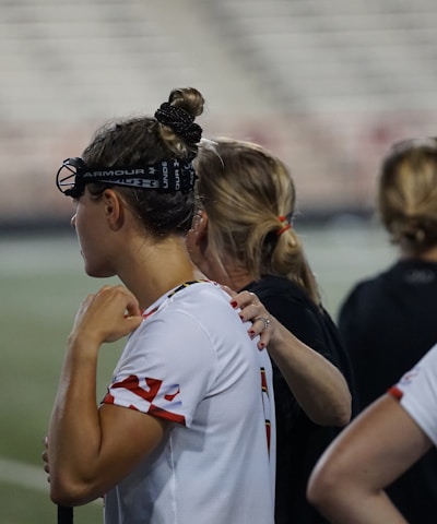 Several people wearing sports attire and protective eyewear are gathered on a sports field. They appear to be engaged in a discussion or briefing, possibly during a break from a game or practice. The background shows a blurred stadium seating, indicating an outdoor setting.