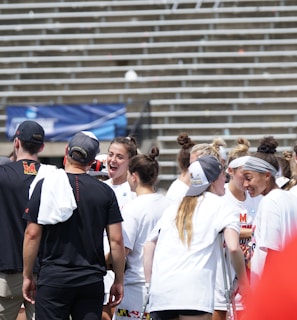 A group of athletes in white shirts with black and red accents are gathered together, engaged in conversation and smiling. They appear to be on a sports field with empty bleachers in the background. The setting is outdoors, and it seems sunny. Some individuals are wearing caps or headbands.