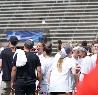 A group of athletes in white shirts with black and red accents are gathered together, engaged in conversation and smiling. They appear to be on a sports field with empty bleachers in the background. The setting is outdoors, and it seems sunny. Some individuals are wearing caps or headbands.