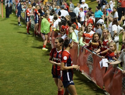 The image features a lively gathering of young soccer players and fans next to a field. Several female athletes in red and blue uniforms interact with an enthusiastic crowd standing behind a barrier. Some people in the crowd are holding objects, like pens and paper, possibly for autographs.